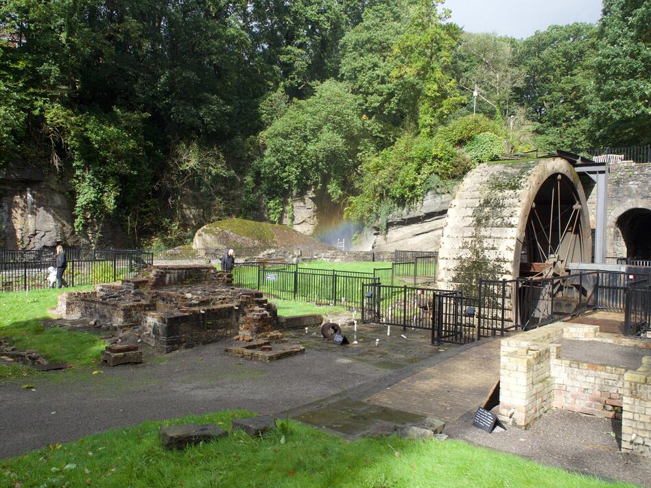 A picture with a waterwheel to the right, a few partially collapsed brick structures, and a rainbow on the waterfall in the background.